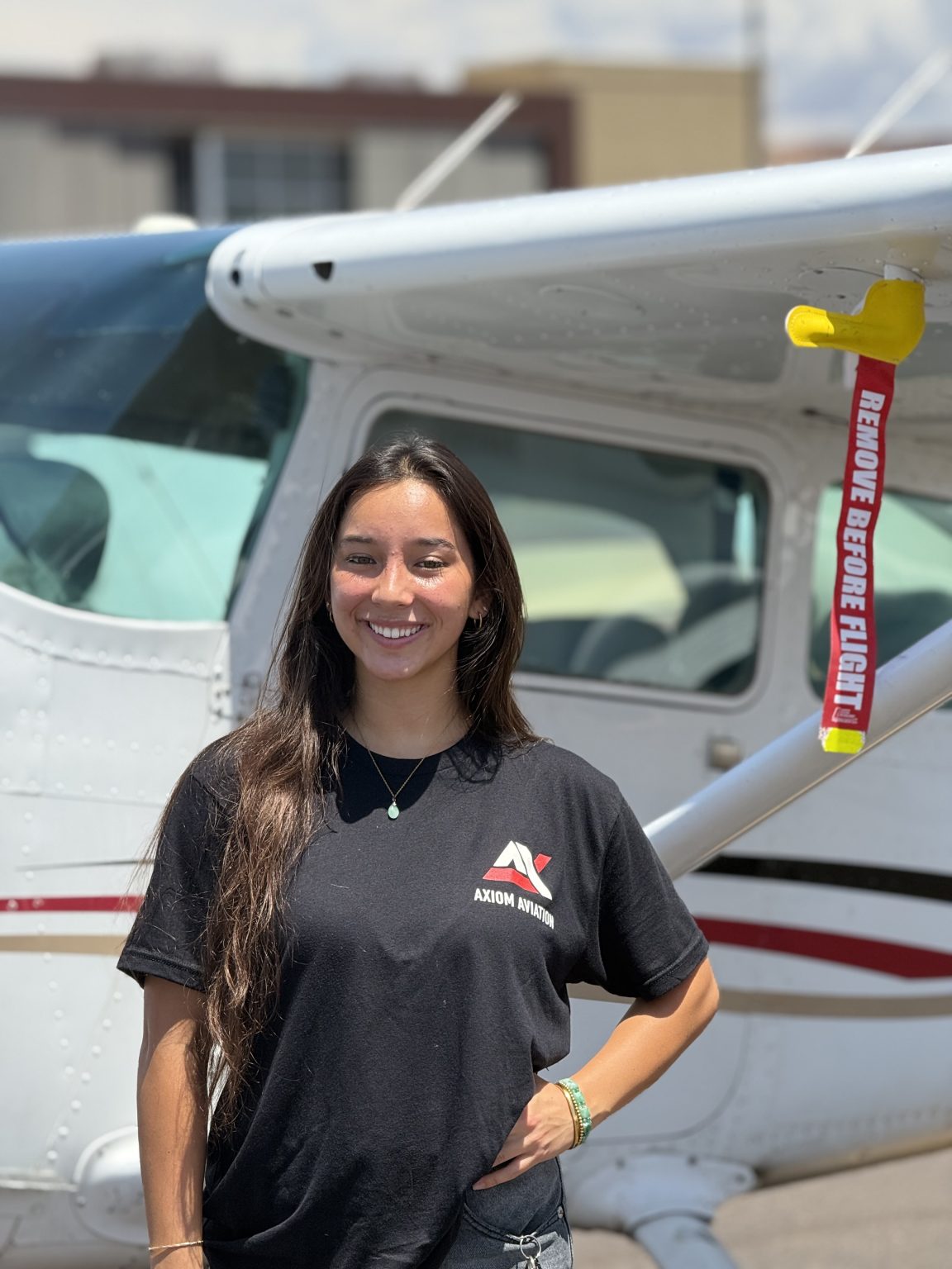 Smiling female student pilot in an Axiom Aviation shirt standing beside a high-wing Cessna trainer on the ramp