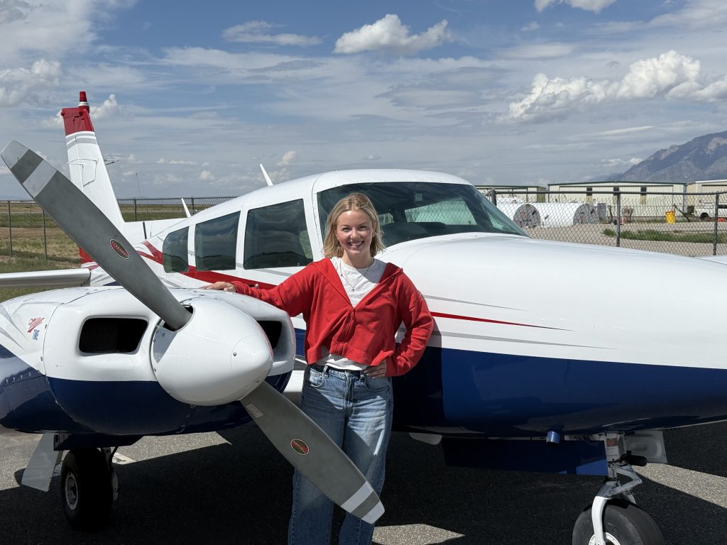 Female pilot standing at the propeller of a red, white and blue twin-engine airplane with mountains behind